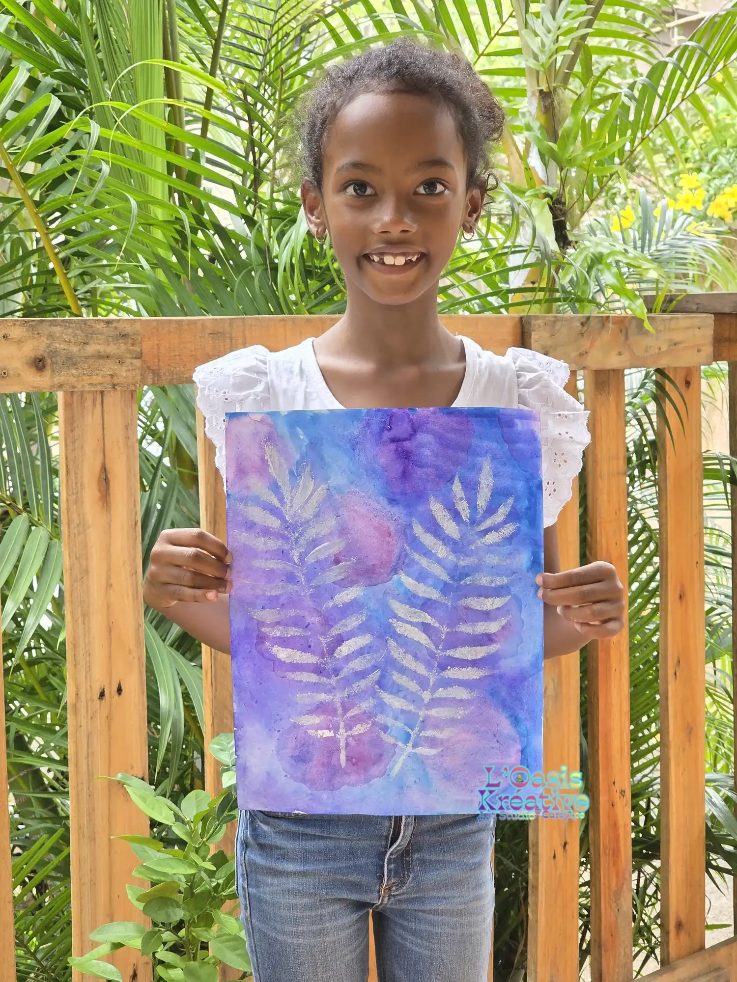 Une jeune fille sourit fièrement en présentant sa peinture de feuilles tropicales argentées sur fond aquarelle violet et bleu