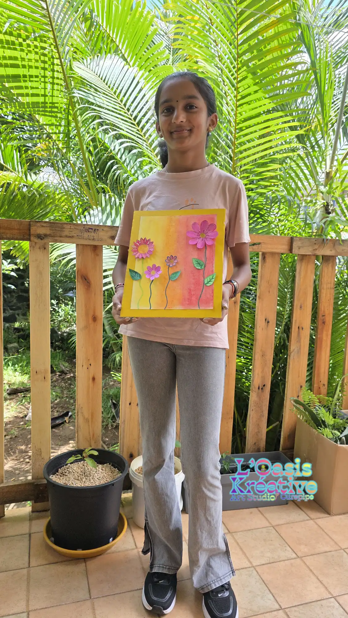 Une jeune fille debout présente fièrement son tableau en relief composé de fleurs roses sur un fond dégradé jaune et orangé.