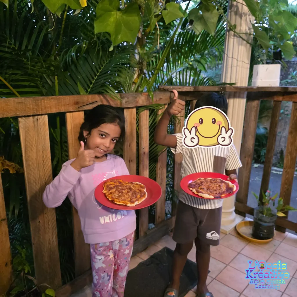 Deux enfants souriants sur une terrasse en bois, tenant chacun une pizza artisanale sur une assiette rouge et faisant un pouce levé, lors d'un atelier à L'Oasis Kréative.