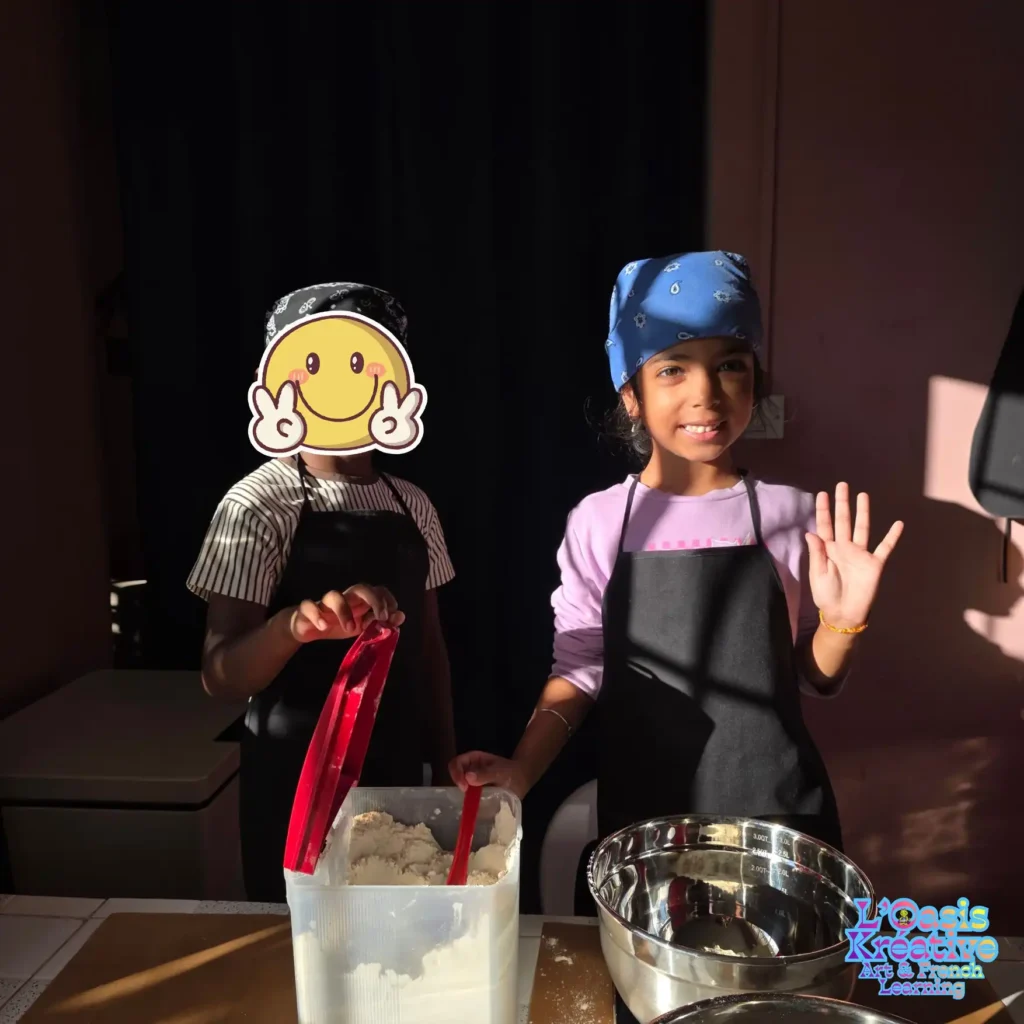 Deux enfants portant des tabliers noirs participent à un atelier de cuisine créative à l'Oasis Kréative avec Zazie Jam, manipulant de la farine dans une ambiance lumineuse.