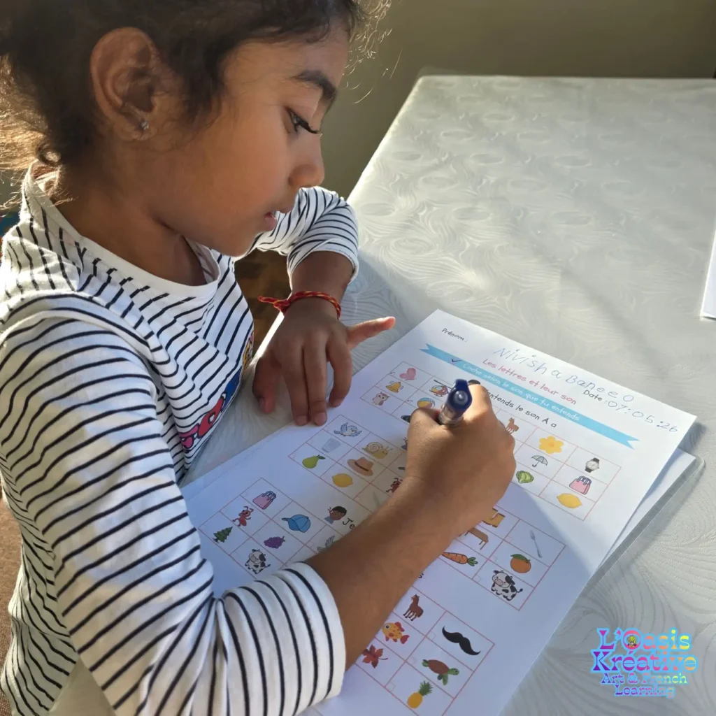 Une petite fille concentrée remplit une fiche d'exercices ludiques sur les sons et les lettres en français lors d'un atelier à L'Oasis Kréative.