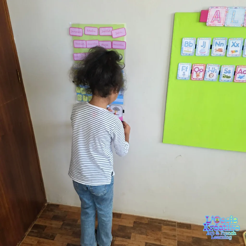 Une jeune fille de dos manipulant un calendrier interactif des mois et de la météo lors d'un cours d'éveil au français à L'Oasis Kréative.