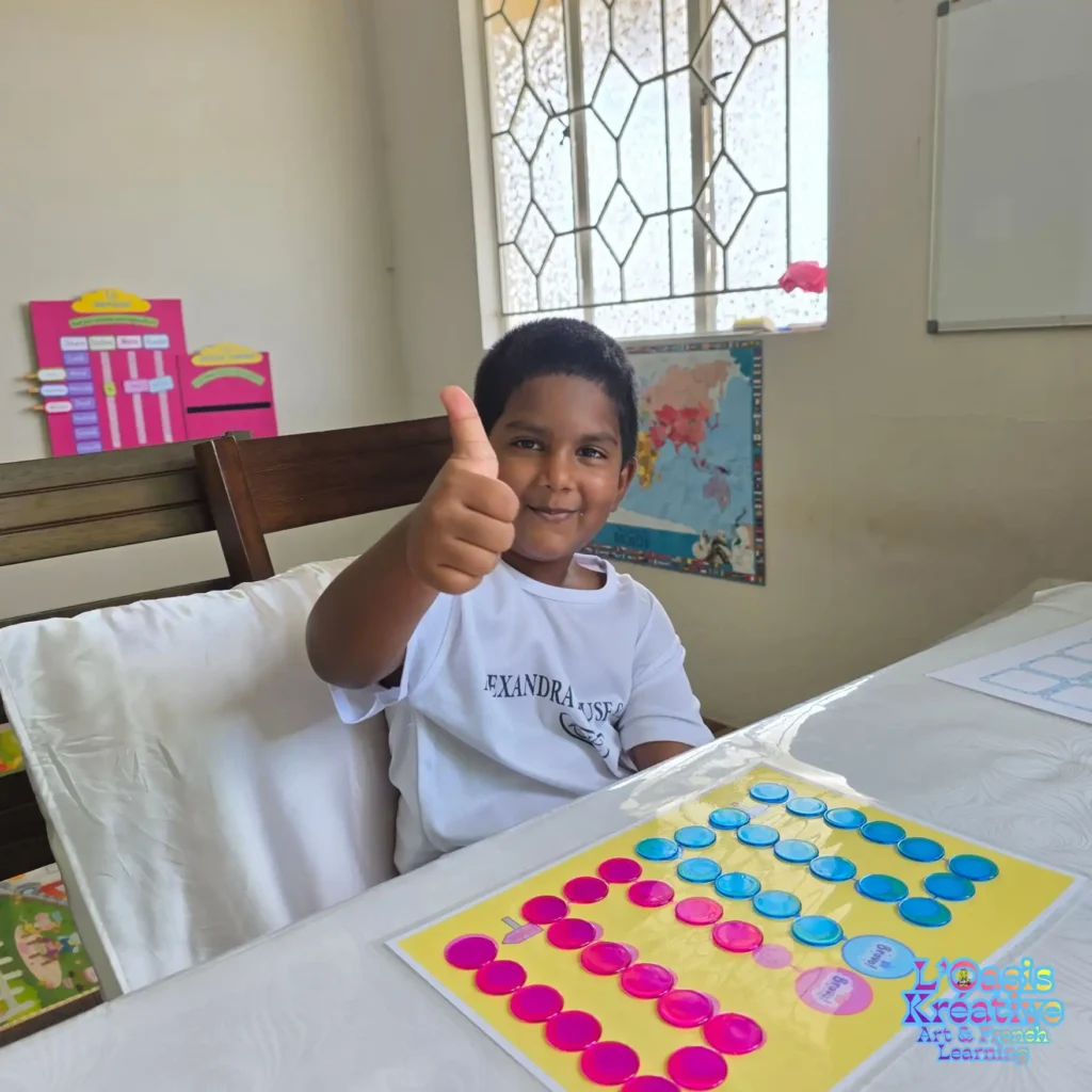 Un jeune garçon souriant fait un pouce levé devant un jeu éducatif de jetons colorés lors d'un cours d'éveil au français à L'Oasis Kréative.