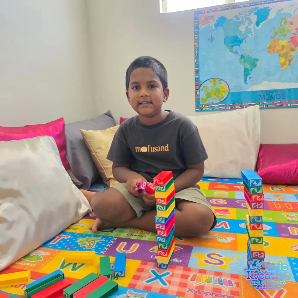 Jeune enfant assis sur un tapis alphabet coloré, empilant des blocs de construction numérotés lors d'une session d'éveil au français à l'Oasis Kréative.