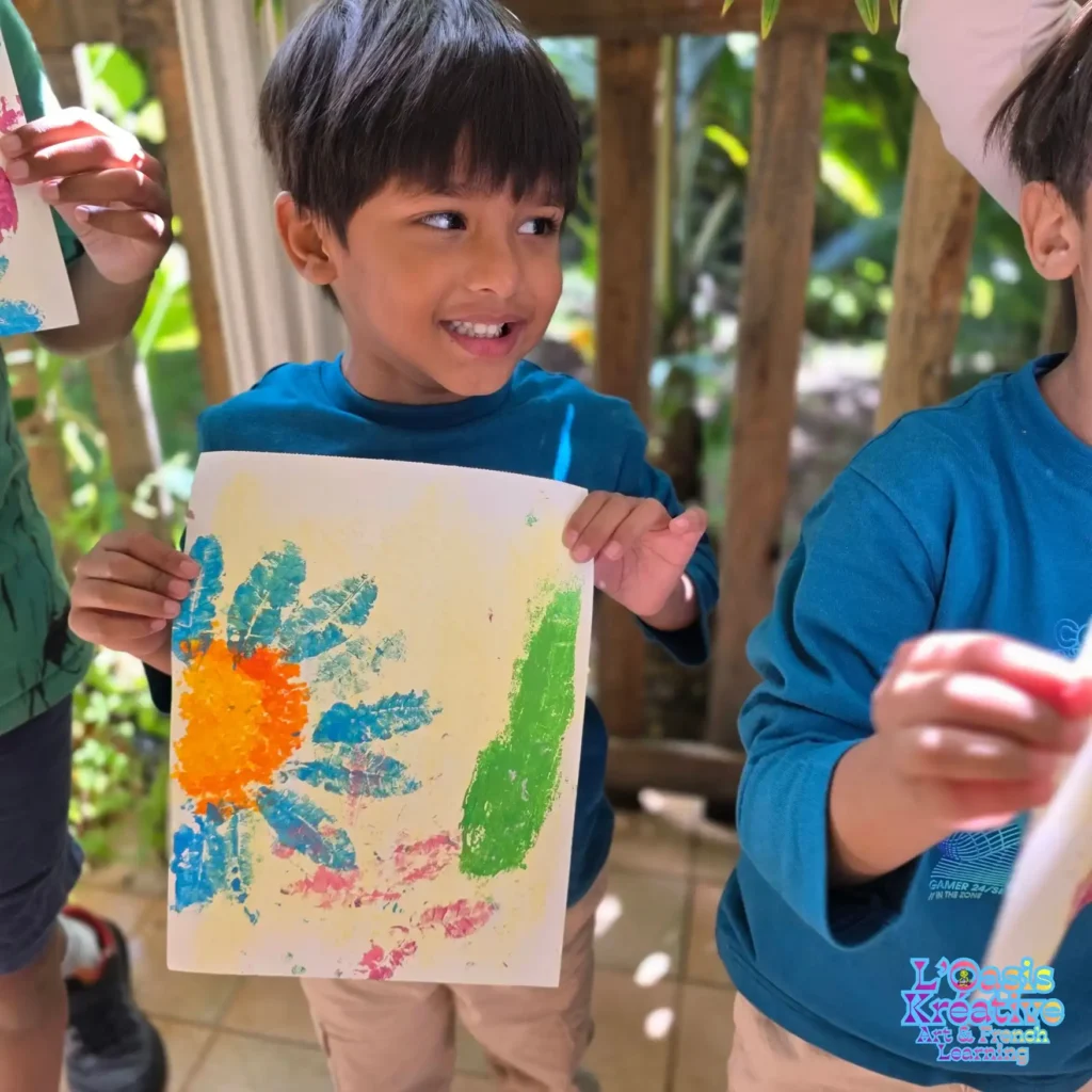 Jeune enfant montrant un tableau coloré réalisé en impressions végétales avec des feuilles et de la peinture lors d’un atelier créatif avec Zazie Jam à L’Oasis Kréative – Art & French Learning à Curepipe.