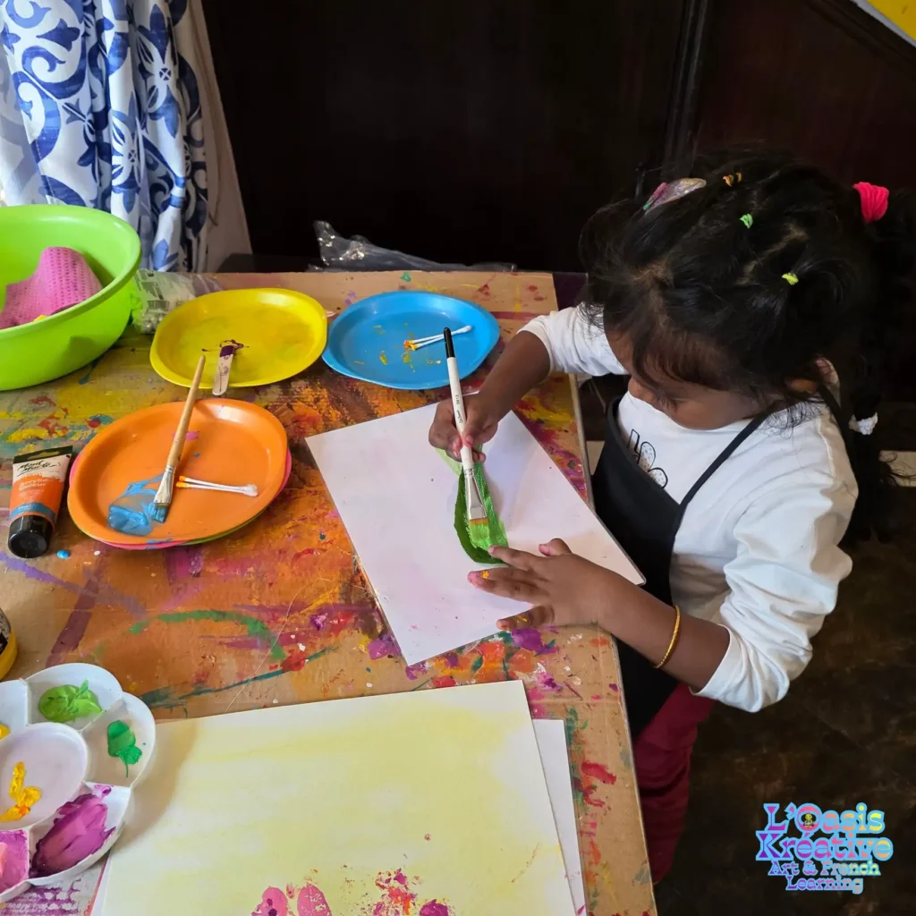Une petite fille peint soigneusement une feuille d'arbre verte avec un pinceau lors d'un atelier d'impressions végétales à L'Oasis Kréative à Curepipe. Option 2 (Plus courte) : Enfant peignant une feuille verte pour un atelier de tableaux en impressions végétales.