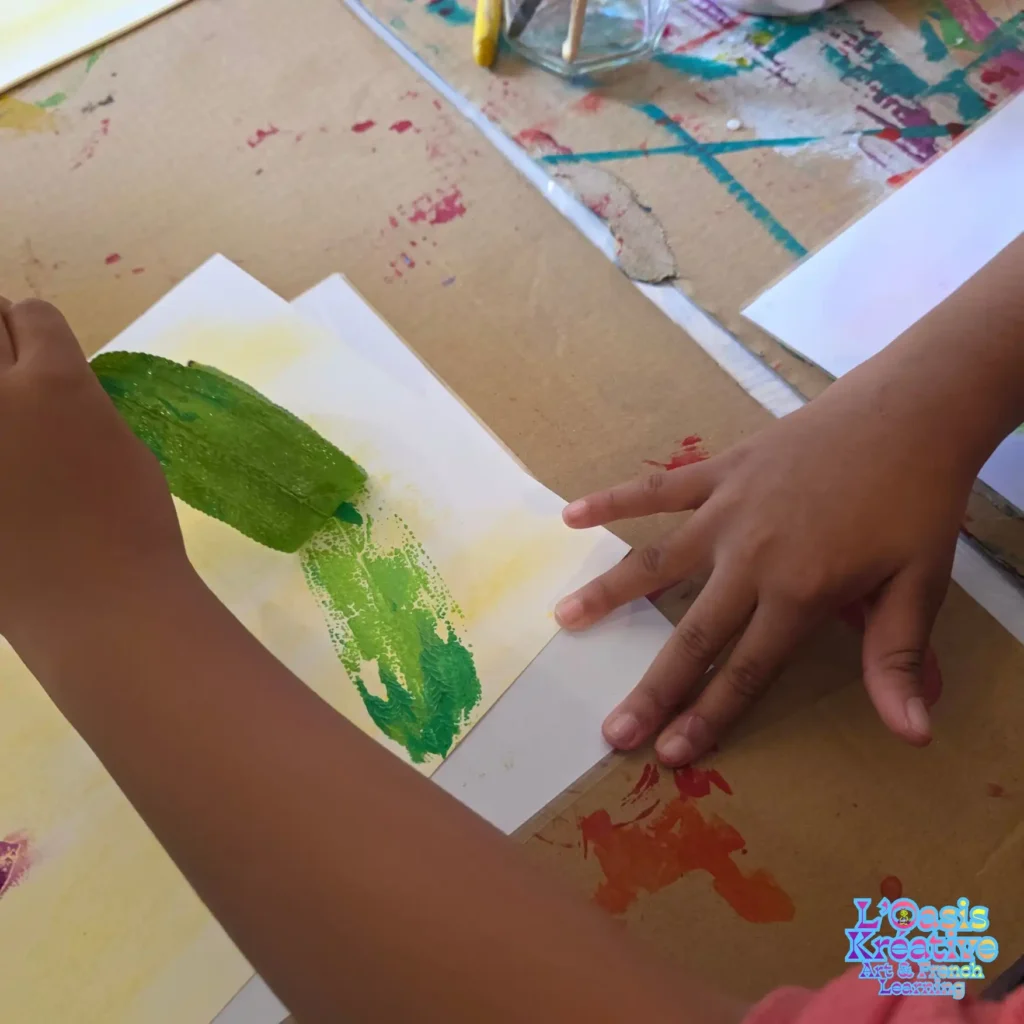 Gros plan sur les mains d'un enfant appliquant une feuille peinte en vert pour créer une empreinte végétale à L'Oasis Kréative, Curepipe.