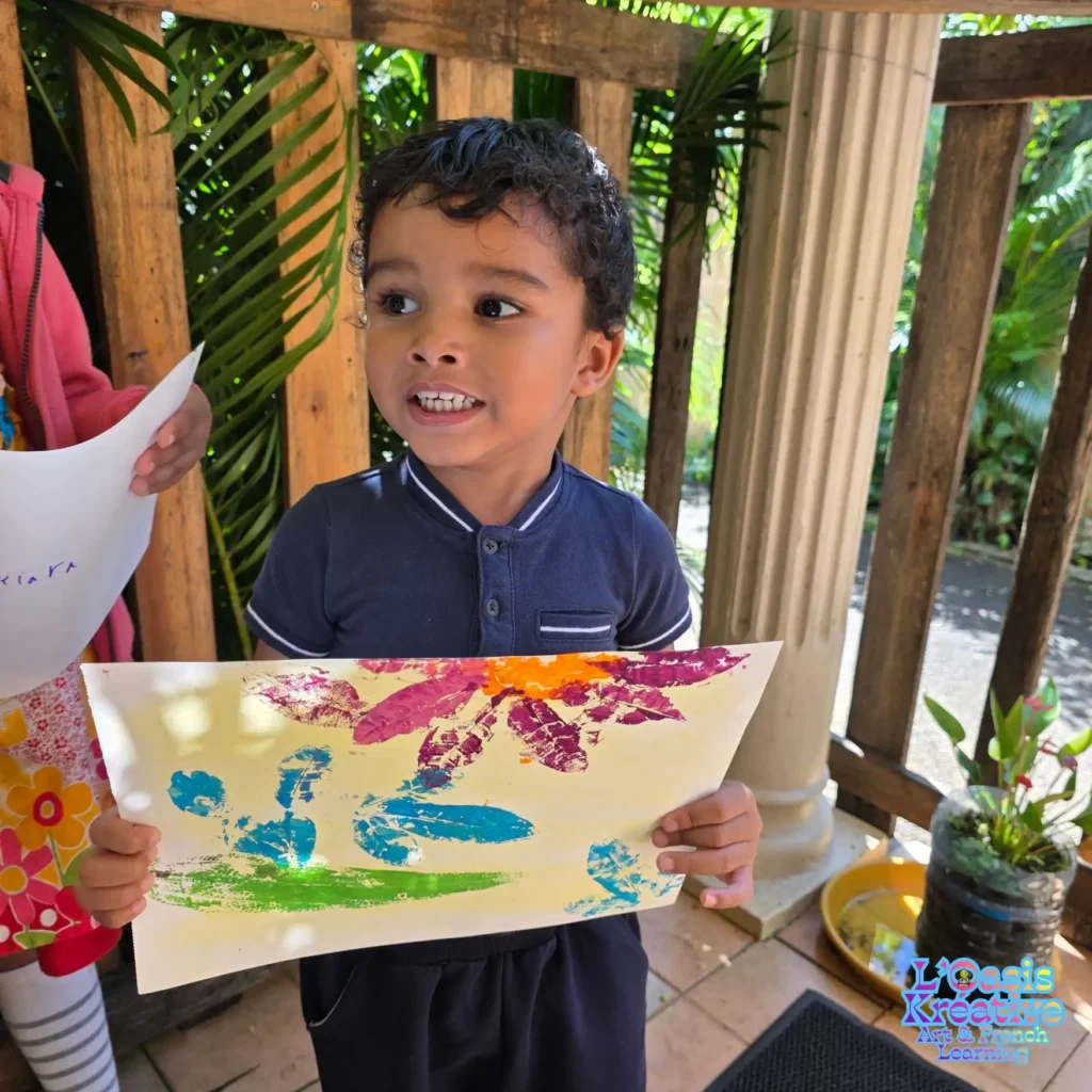 Jeune enfant montrant un tableau coloré réalisé en impressions végétales avec des feuilles et de la peinture lors d’un atelier créatif animé par Zazie Jam à L’Oasis Kréative – Art & French Learning à Curepipe.