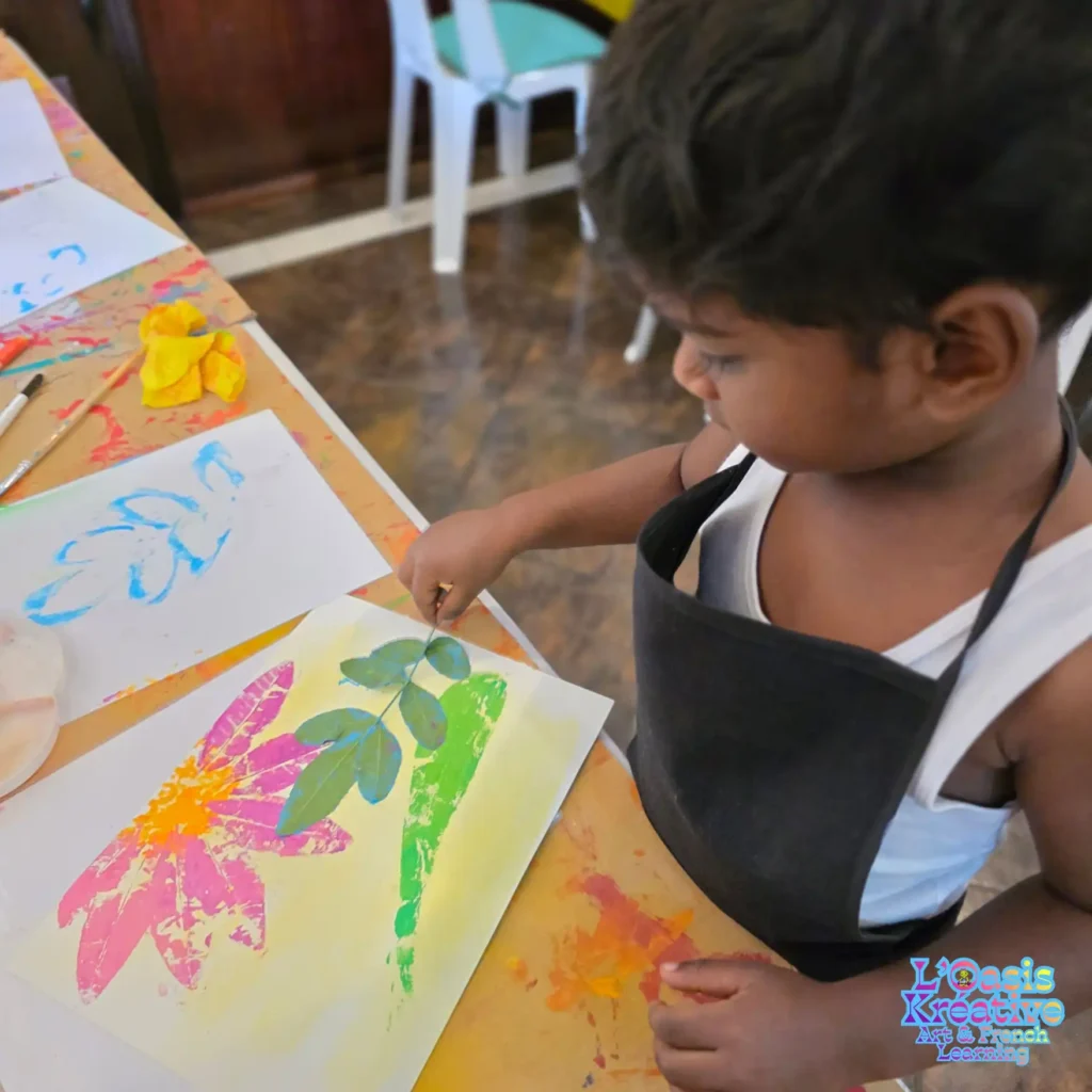 Un jeune garçon, portant un tablier noir, place délicatement une feuille peinte sur son dessin pour créer une empreinte végétale à l'Oasis Kréative, à Curepipe. Sur le papier se trouvent déjà des empreintes de fleurs roses et d'herbes vertes.