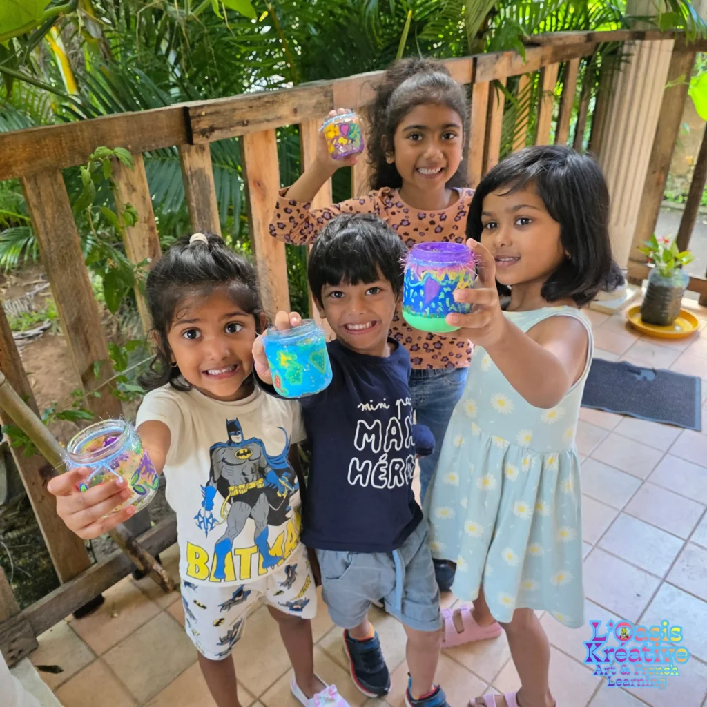 Texte alt :

Quatre enfants souriants brandissant fièrement leurs pots en verre peints à la main lors d'un atelier créatif en plein air à L'Oasis Kréative à Maurice.