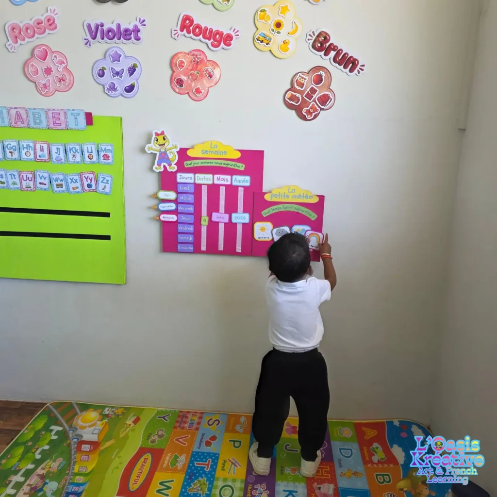 Jeune enfant participant au rituel de la météo et du calendrier en français lors d'un atelier à L'Oasis Kréative à Curepipe, Île Maurice. Le mur est décoré d'affichages pédagogiques colorés sur les couleurs et l'alphabet.
