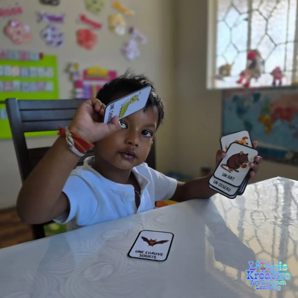 Enfant participant à un atelier d'immersion en français à L'Oasis Kréative à Curepipe, Maurice, jouant avec des cartes de vocabulaire illustrées sur les animaux ("une chauve-souris", "un rat", "une chenille").